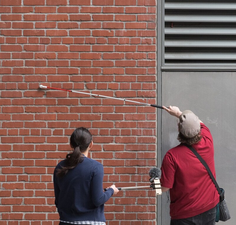Andy Slater runs a long white cane along an exterior wall of the CJM. A person with hair in a ponytail holds a microphone out to capture the sound.